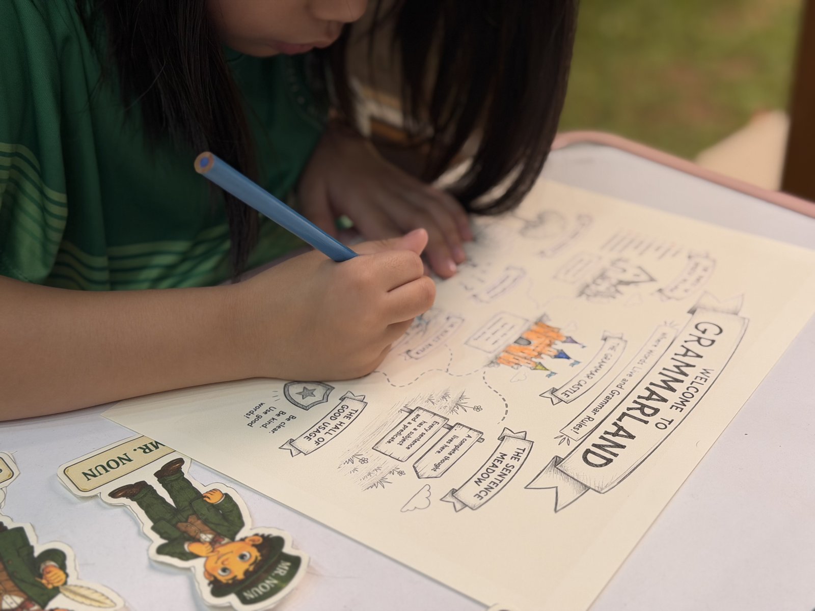 A young Filipino homeschooler coloring the Welcome to Grammarland map with a Mr. Noun character cutout on the table