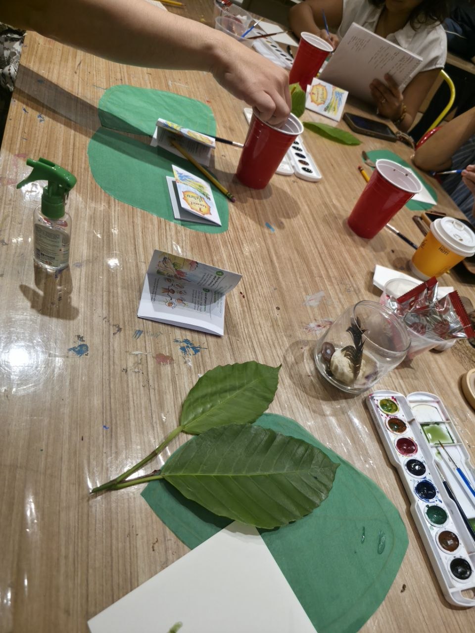 A workshop table with fresh leaves, watercolour palettes, brushes, cups, and booklets during a nature-journaling session
